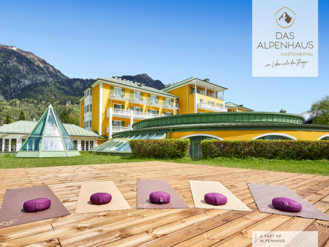 Yoga-Setup im Freien mit Matten und Kissen auf einem Holzdeck gegenüber einem modernen Gebäude. Das Gebäude hat gelbe Wände und grüne Dächer. Im Hintergrund sind Berge unter einem klaren blauen Himmel zu sehen. Auf dem Schild steht „Das Alpenhaus Gasteinertal“.