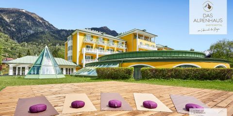 Vor einem leuchtend gelben Hotel steht ein sonnenbeschienenes Outdoor-Yogadeck mit Matten und violetten Nackenrollen. Im Hintergrund sind Berge und ein klarer blauer Himmel zu sehen. In der Nähe ist ein Wintergarten in Form einer Glaspyramide zu sehen.