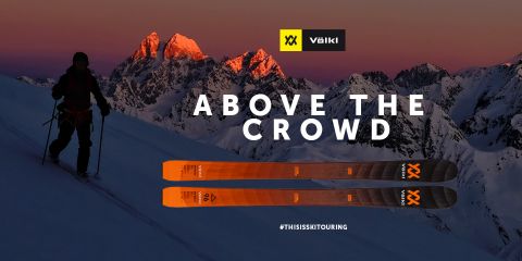 Ein Skifahrer steht in der Abenddämmerung auf einem schneebedeckten Berg und hebt sich als Silhouette vom Himmel ab. Im Hintergrund sind die leuchtenden Gipfel zu sehen. Im Vordergrund sind orangefarbene Völkl-Ski zu sehen. Der Text lautet „Above the Crowd“ mit den Hashtags #THISISSKTOURING.