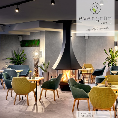 Moderne Lounge mit zentralem, kegelförmigem Kamin. Der Raum ist mit grünen und gelben Stühlen, kleinen Holztischen und üppigen Pflanzen ausgestattet. Der Raum ist durch Spots an der Decke gut beleuchtet. In der Ecke ist das Branding für „ever.grün KAPRUN“ sichtbar.