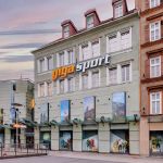 Außenansicht eines Gigasport-Geschäfts in einer städtischen Umgebung. Das Gebäude ist drei Stockwerke hoch und hat große Schaufenster, in denen Outdoor- und Sportartikel angeboten werden. Das Logo des Geschäfts ist prominent über dem Eingang zu sehen. Der Himmel ist in der Abenddämmerung.