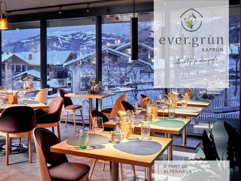 Modernes Restaurant-Interieur mit hübsch gedeckten Tischen, jeder mit Weingläsern und grünen Dekorationen. Große Fenster geben den Blick auf eine verschneite Bergdorflandschaft frei. Ein Logo lautet „ever.grün KAPRUN“ mit einem Slogan und einem Hinweis, dass es Teil von „ALPENHAUS“ ist.