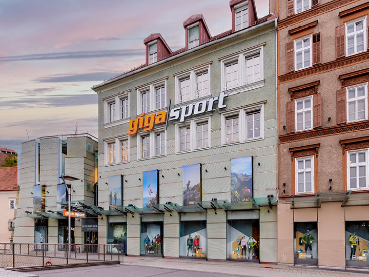 Straßenansicht eines Gigasport-Geschäfts mit hellgrüner Fassade und großen Fenstern, in denen Outdoor-Sportartikel ausgestellt sind. Der Eingang hat ein modernes Glasdach und das Gebäude grenzt an ein braunes Backsteingebäude. Der Himmel ist bewölkt und lässt einen Sonnenuntergang erahnen.