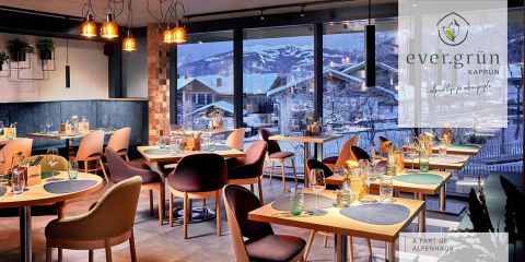 Ein modernes, elegantes Restaurant mit gedeckten Tischen. Große Fenster geben den Blick auf ein verschneites Bergdorf frei. Warme Beleuchtung und stilvolles Dekor sorgen für eine gemütliche Atmosphäre. Ein Logo in der Ecke lautet „ever.grün Kaprun, ein Teil von Alpenhaus“.