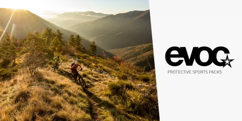 Zwei Mountainbiker fahren einen sonnenbeschienenen Pfad entlang, der von üppigem Grün und sanften Hügeln umgeben ist. Die Sonne wirft einen warmen Schein über die Landschaft. Auf der rechten Seite des Bildes steht ein Logo mit der Aufschrift „EVOC: Protective Sports Packs“.