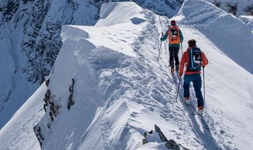 Zwei Menschen mit Rucksäcken und Winterkleidung wandern einen schmalen, schneebedeckten Bergkamm entlang. Sie stützen sich mit Stöcken ab, umgeben von steilen, schneebedeckten Gipfeln unter klarem Himmel.
