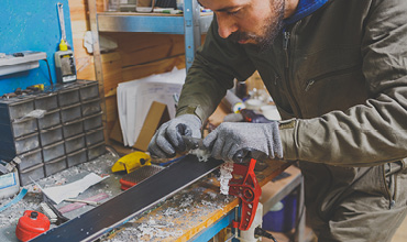 Eine Person in Jacke und Handschuhen spannt in einer Werkstatt einen Ski. Sie benutzt ein Werkzeug, um den Belag abzuschaben. Auf der Werkbank liegen verschiedene Werkzeuge und Materialien verstreut. Im Hintergrund sind Regale mit zusätzlicher Ausrüstung zu sehen.