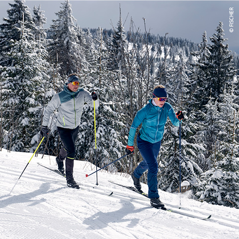 Zwei Personen laufen auf einer verschneiten Loipe durch eine Waldlandschaft. Sie tragen Winterkleidung, darunter Jacken und Sonnenbrillen, und bewegen sich energisch unter einem teilweise bewölkten Himmel. Sie sind von schneebedeckten Bäumen umgeben.