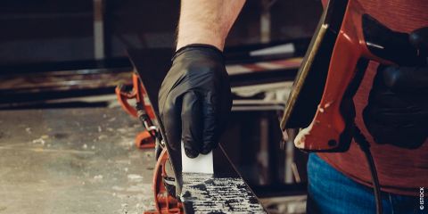 Eine Person mit schwarzen Handschuhen benutzt ein Werkzeug zum Wachsen eines Snowboards, das in einer Klemme befestigt ist. Der Arbeitsbereich mit verschiedenen Geräten ist zu sehen, wobei der Fokus auf dem Auftragen von Wachs auf die Oberfläche des Boards liegt.