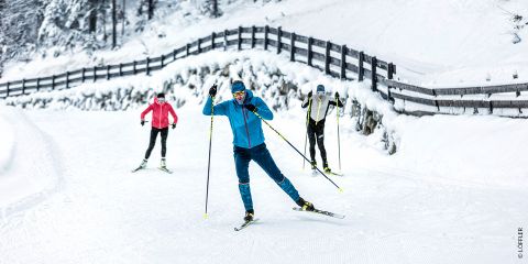Drei Personen laufen auf einer verschneiten Loipe Ski. Der Skifahrer vorn trägt ein blaues Outfit, zwei weitere folgen ihm, einer in Rosa, der andere in Grau. Schneebedeckte Bäume und ein Holzzaun säumen den Weg.