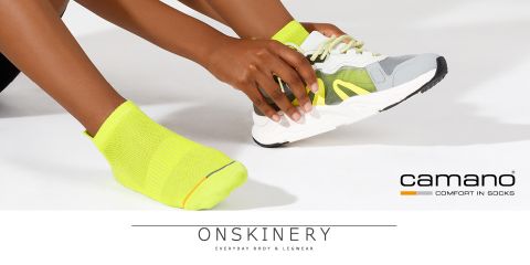 Person zieht einen weißen Sneaker mit grauen und fluoreszierenden gelben Akzenten über einer leuchtend gelben Socke an. Ein anderer Fuß zeigt dieselbe gelbe Socke. Der Text lautet „Camano Comfort in Socks“ und „Onskinery Everyday Body & Legwear“.