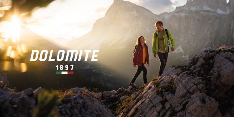 Ein Mann und eine Frau in Wanderkleidung gehen bei Sonnenaufgang oder Sonnenuntergang auf felsigem Gelände spazieren, mit Bergen im Hintergrund. Der Text lautet „DOLOMITE 1897“ mit einer kleinen italienischen Flagge darunter.