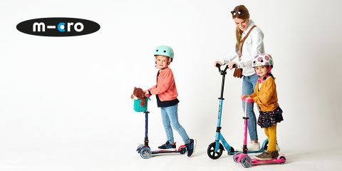 Eine Frau und zwei Kinder fahren bunte Roller. Die Frau trägt eine Handtasche, während der Junge einen Teddybären in einem Korb auf seinem Roller hält. Das Mädchen trägt einen Helm mit Mustern. Alle lächeln vor einem weißen Hintergrund mit einem „Micro“-Logo.
