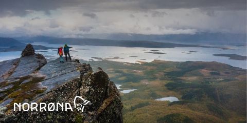 Zwei Wanderer stehen auf einer Felsklippe und blicken unter einem bewölkten Himmel auf eine weite Landschaft aus Seen und Wäldern. In der unteren linken Ecke ist ein Logo mit „Norrøna“ zu sehen.