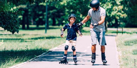 Ein Mann und ein kleiner Junge fahren auf Rollschuhen auf einem glatten Weg in einem sonnigen Park. Beide tragen Helme und Schutzausrüstung. Der Mann stützt den Jungen mit einer führenden Hand, während sie zwischen grünen Bäumen und Gras dahingleiten.