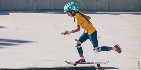 Ein junges Mädchen mit blaugrünem Helm, gelbem Hemd, Jeans und Schutzkleidung fährt in einem Skatepark Skateboard. Sie ist in Bewegung, stößt sich mit einem Fuß ab und balanciert mit dem anderen auf dem Brett. Der Hintergrund ist eine Betonfläche.