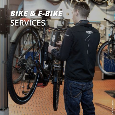 Eine Person in einer schwarzen Jacke führt Wartungsarbeiten an einem Fahrrad durch, das in einem vertikalen Ständer in einem Fahrradladen befestigt ist. In der oberen linken Ecke wird der Text „BIKE & E-BIKE SERVICES“ angezeigt. Im Hintergrund sind verschiedene Fahrradteile zu sehen.