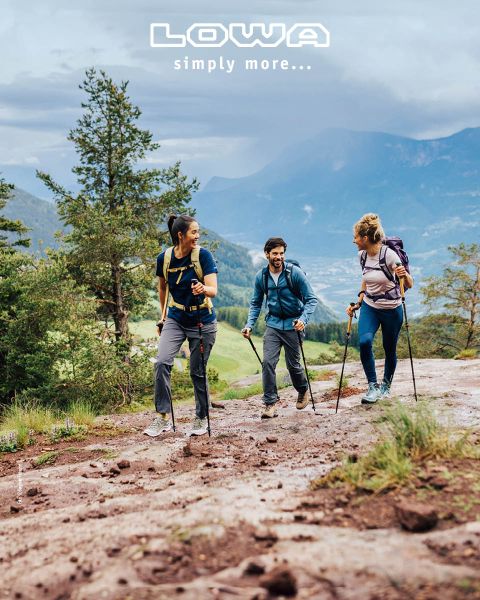 Drei Personen wandern mit Trekkingstöcken und Outdoor-Ausrüstung auf einem malerischen Bergpfad. Im Hintergrund sind Bäume und Berge zu sehen. Der Himmel ist bewölkt. Oben steht der Schriftzug „LOWA simply more…“.