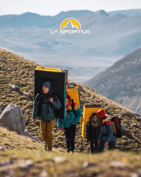 Vier Personen in Outdoor-Ausrüstung und mit Bouldermatten auf dem Rücken wandern einen grasbewachsenen Bergpfad hinauf. Über ihnen ist das Logo von La Sportiva am Himmel zu sehen. Im Hintergrund sind schroffe Berggipfel zu sehen.