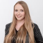 Frau mit langen blonden Haaren, trägt eine schwarze Lederjacke und ein schwarzes Oberteil und lächelt vor einem schlichten weißen Hintergrund.