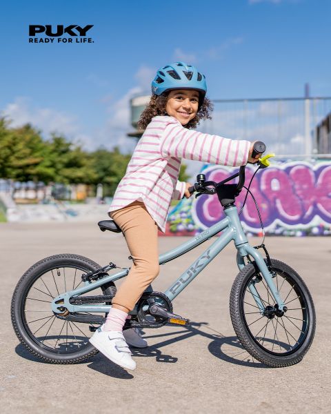 Ein junges Mädchen mit blauem Helm und gestreiftem Shirt fährt an einem sonnigen Tag auf einem hellblauen Fahrrad. Lächelnd fährt sie an bunten Graffitis in einer urbanen Umgebung vorbei. Das PUKY-Logo und der Slogan „Ready for life“ sind oben links zu sehen.