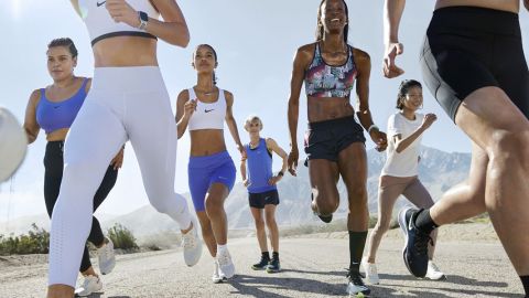 Eine bunt gemischte Gruppe joggt an einem sonnigen Tag im Freien. Sie tragen Sportkleidung, darunter Shorts, Tanktops und Turnschuhe. Im Hintergrund ist eine Berglandschaft zu sehen. Sie wirken voller Energie und konzentrieren sich auf ihren Lauf.