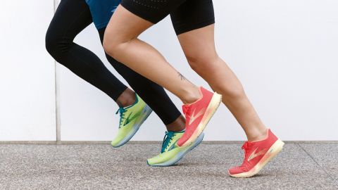 Zwei Menschen laufen und präsentieren sportliche Turnschuhe. Einer trägt limettengrüne Schuhe mit blauen Akzenten und der andere korallenfarbene Schuhe mit beigen Sohlen. Beide sind in Bewegung vor einer weißen Wand und einem grauen Gehweghintergrund.