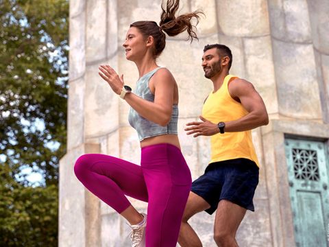 Zwei Menschen machen Sport im Freien. Eine Frau in grauem Sport-BH und rosa Leggings joggt energisch. Hinter ihr läuft ein Mann in gelbem Tanktop und schwarzen Shorts. Sie wirken konzentriert und stehen vor einer Steinstruktur.