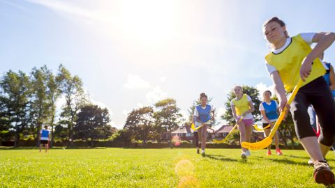 Eine Gruppe Kinder spielt Feldhockey auf einem Rasenplatz. Sie tragen Sportkleidung und halten gelbe Hockeyschläger in der Hand. Die Sonne scheint hell, im Hintergrund sind Bäume und Häuser unter einem klaren Himmel zu sehen.