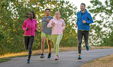 Vier Personen joggen auf einem gepflasterten Weg in einem Park. Sie tragen farbenfrohe Sportkleidung und scheinen den Lauf zu genießen. Im Hintergrund sind grüne Bäume und Gras unter einem klaren Himmel zu sehen.