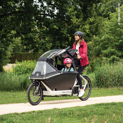 Eine Frau mit roter Jacke und schwarzem Helm fährt mit einem elektrischen Lastenfahrrad auf einem Weg durch einen Park. Ein Kind mit rosa Helm sitzt im vorderen Bereich des Fahrrads, der von einem Baldachin bedeckt ist. Im Hintergrund sind Bäume und Gras zu sehen.