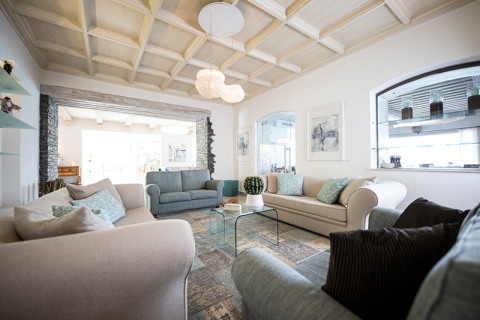 Ein gemütliches Wohnzimmer mit zwei beigen Sofas und einer blauen Couch, die um einen Couchtisch aus Glas angeordnet sind. Der Raum verfügt über eine Kassettendecke, moderne Leuchten und dekorative Regale, die eine warme, einladende Atmosphäre schaffen.