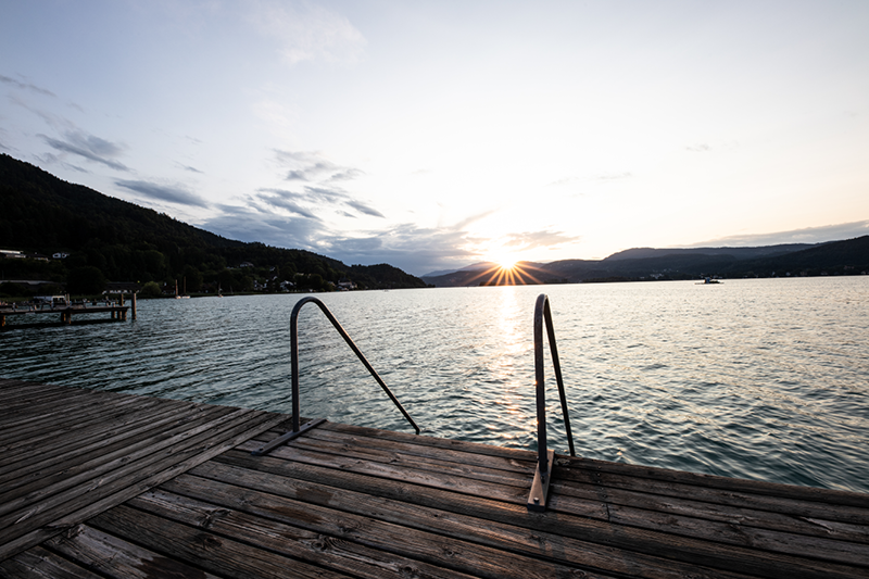 Sonnenuntergang über einem ruhigen See, mit einem Holzsteg im Vordergrund und einer Leiter, die zum Wasser führt. Die Sonne versinkt hinter fernen Hügeln und wirft einen sanften Schein auf die vereinzelten Wolken am Himmel.