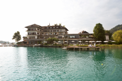 Ein mehrstöckiges Hotel am Seeufer mit modernem Design, mit Balkonen und einem Steg mit Liegestühlen und Sonnenschirmen. Im Vordergrund ist das klare, türkisfarbene Wasser des Sees zu sehen, im Hintergrund sind Bäume und Hügel zu sehen.