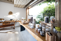 Ein helles Frühstücksbuffet mit verschiedenen Müslis in Gläsern, einer Kaffeemaschine und Gebäck auf Holztischen. Sonnenlicht strömt durch große Fenster, erhellt den Raum und bietet einen Blick ins Grüne draußen. Steinböden ergänzen die rustikale Einrichtung.