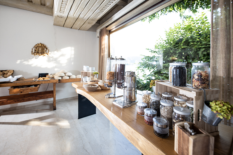 Ein helles Frühstücksbuffet mit verschiedenen Müslis in Gläsern, einer Kaffeemaschine und Gebäck auf Holztischen. Sonnenlicht strömt durch große Fenster, erhellt den Raum und bietet einen Blick ins Grüne draußen. Steinböden ergänzen die rustikale Einrichtung.