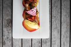 Ein rechteckiger weißer Teller mit farbenfroher, kunstvoller Präsentation der Speisen. Er enthält dünn geschnittenes Gemüse, eine Scheibe rosa-weißer Rüben und eine Kugel gelbe Mousse, angerichtet auf einer rustikalen Holzoberfläche.