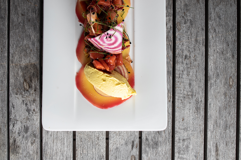 Ein rechteckiger weißer Teller mit farbenfroher, kunstvoller Präsentation der Speisen. Er enthält dünn geschnittenes Gemüse, eine Scheibe rosa-weißer Rüben und eine Kugel gelbe Mousse, angerichtet auf einer rustikalen Holzoberfläche.