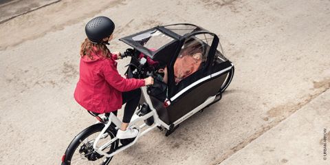 Eine Person mit roter Jacke und schwarzem Helm fährt mit einem elektrischen Lastenfahrrad auf einer asphaltierten Straße. Das Fahrrad verfügt über einen vorderen Laderaum mit Regenschutz, in dem ein Kind sitzt.