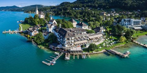Eine malerische Luftaufnahme eines Dorfes am Seeufer mit einer Kirche, Häusern und einem Hotel entlang der Uferlinie. Das türkisfarbene Wasser umgibt das Land, mit üppigen grünen Hügeln im Hintergrund und kleinen Anlegestellen, die in den See hineinragen.