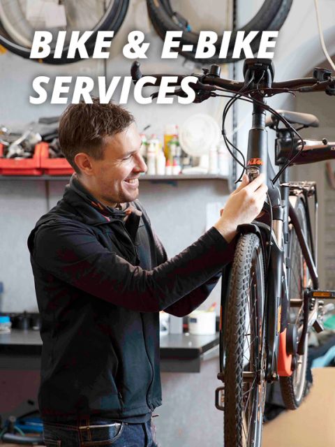 Eine Person in schwarzer Jacke lächelt und stellt in einer Werkstatt das Vorderrad eines Fahrrads ein, das auf einem Ständer montiert ist. Oben im Bild ist der Text „BIKE & E-BIKE SERVICES“ zu sehen. Im Hintergrund sind Werkzeuge und Geräte zu sehen.