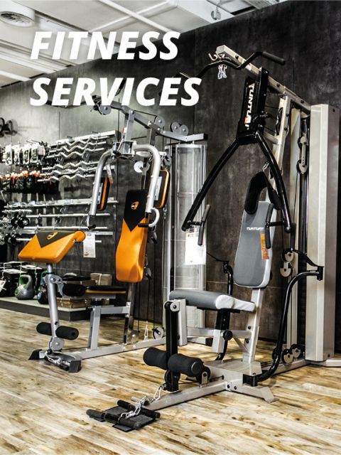 Ein Fitnessstudio-Interieur mit zwei multifunktionalen Trainingsgeräten und verschiedenen Fitnessgeräten im Hintergrund. Oben steht in fetten weißen Buchstaben „Fitness Services“. Der Boden ist aus Holz.
