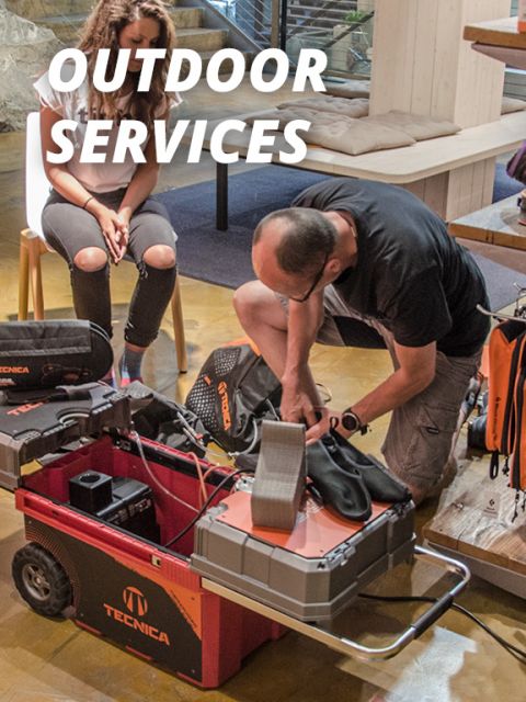 Eine Person kniet neben einem Wagen mit der Aufschrift „TECNICA“ und trägt Stiefel und Ausrüstung. Eine andere Person sitzt daneben. Der Text darüber lautet „OUTDOOR SERVICES“. Die Szene ist in einem Gebäude mit Holzmöbeln und Treppen im Hintergrund zu sehen.