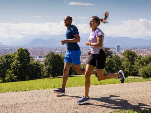 Ein Mann und eine Frau joggen auf einem Pfad in einem Park. Sie tragen Sportkleidung, wobei der Mann ein blaues Oberteil und die Frau ein hellviolettes Oberteil trägt. Im Hintergrund sieht man eine Stadtlandschaft und Berge unter einem teilweise bewölkten Himmel.