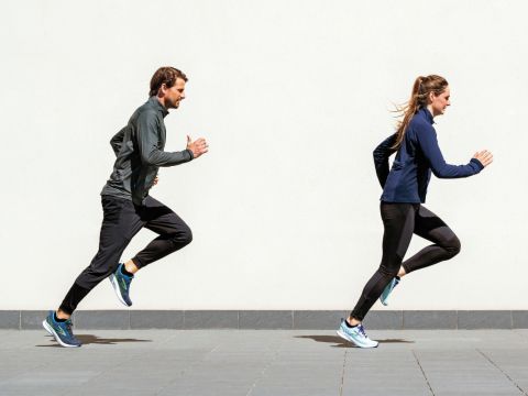 Ein Mann und eine Frau laufen nebeneinander auf einem Bürgersteig an einer schlichten weißen Wand entlang. Beide tragen Sportkleidung, darunter Jacken und Leggings. Sie wirken konzentriert und entschlossen, aufgenommen mitten im Schritt.