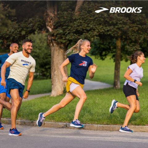Vier Personen joggen auf einem Pfad in einem Park. Sie lächeln und tragen Sportkleidung. Im Hintergrund sind ein großer Baum und Gras zu sehen. Das Markenlogo „Brooks“ erscheint in der oberen rechten Ecke.