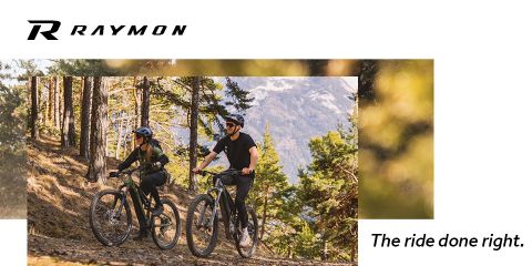 Zwei Personen mit Helmen fahren Mountainbikes auf einem Waldweg. Sie sind von Bäumen umgeben und im Hintergrund sind Berge zu sehen. Der Text lautet „R Raymon“ und „Die Fahrt richtig gemacht“.