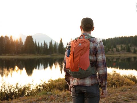Eine Person in kariertem Hemd und Mütze steht bei Sonnenuntergang neben einem spiegelnden See und trägt einen orangefarbenen Rucksack. Im Hintergrund sind Bäume und Berge zu sehen.
