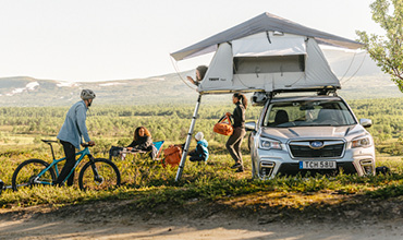 Ein Auto mit einem Dachzelt steht in einer üppigen, grünen Landschaft. Drei Personen bauen ihr Lager auf; zwei stehen neben dem Auto, einer sitzt auf einem Klappstuhl. Ein Radfahrer mit Helm steht daneben. Im Hintergrund sind Hügel zu sehen.