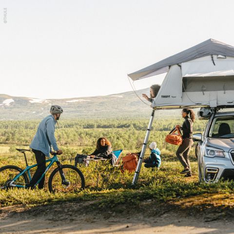 Eine Gruppe von Menschen zeltet in einer malerischen Berggegend. Rechts ist ein Auto mit einem Dachzelt zu sehen. Eine Person ist auf einem Fahrrad unterwegs, zwei Personen sitzen auf Stühlen und eine weitere Person hält eine orangefarbene Tasche. Es ist ein klarer Tag.
